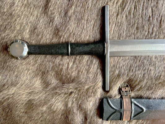 A hand forged carbon steel blade sword with a black grip and cross guard, alongside a wooden scabbard wrapped in leather, displayed on a fur surface.