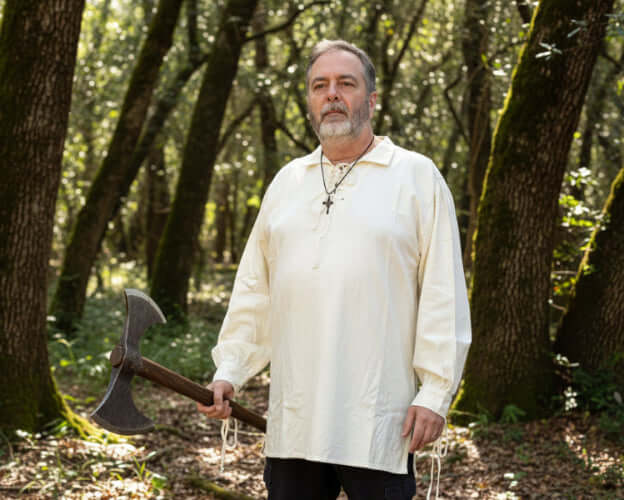 Man wearing a men's medieval cotton shirt holding a large axe, standing in a forest setting.