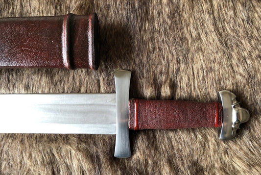 A close-up of a hand forged Viking sword with a leather grip and a metal guard, lying on a fur surface. A wood scabbard with leather fittings is also visible.
