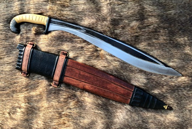 An Iberian Falcata sword with a hand forged carbon steel blade, white wood handle, and a full tang. The sword is accompanied by a wood and leather scabbard.