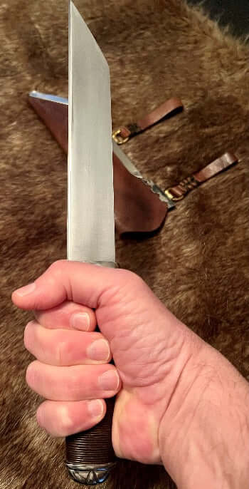 Hjalmar Viking Seax Knife held in hand with leather sheath in background, showcasing its craftsmanship and design.