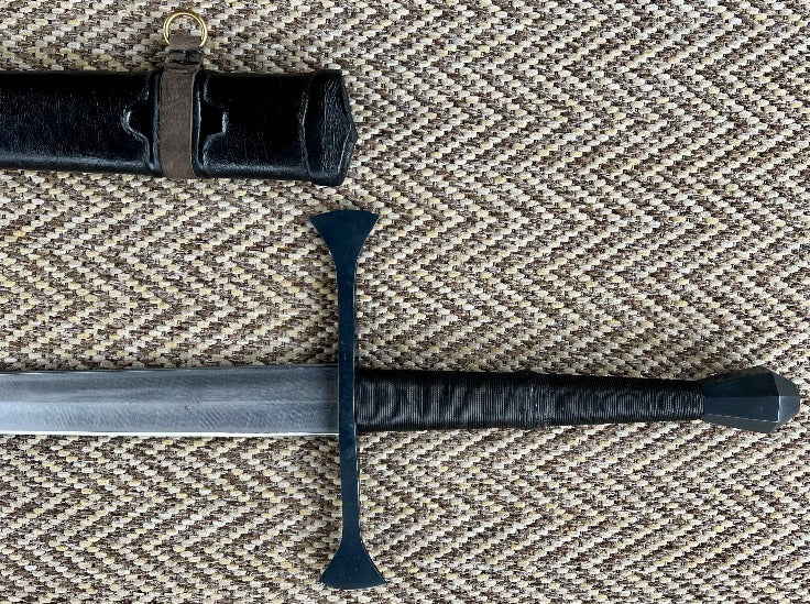 A close-up of a hand forged carbon steel blade with a diamond cross-section, showing the texture and tapered blade. The blade is accompanied by a wooden scabbard wrapped in leather.