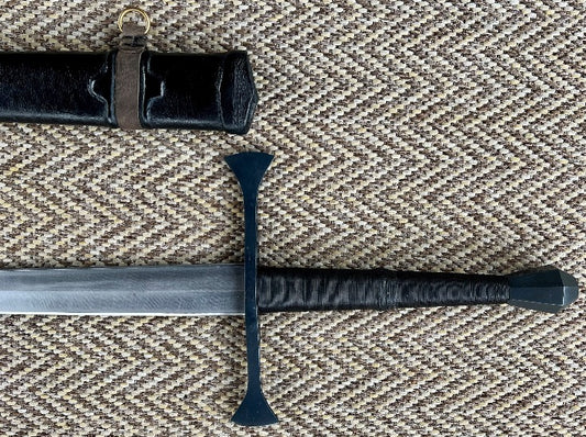 A close-up of a hand forged carbon steel blade with a diamond cross-section, showing the texture and tapered blade. The blade is accompanied by a wooden scabbard wrapped in leather.