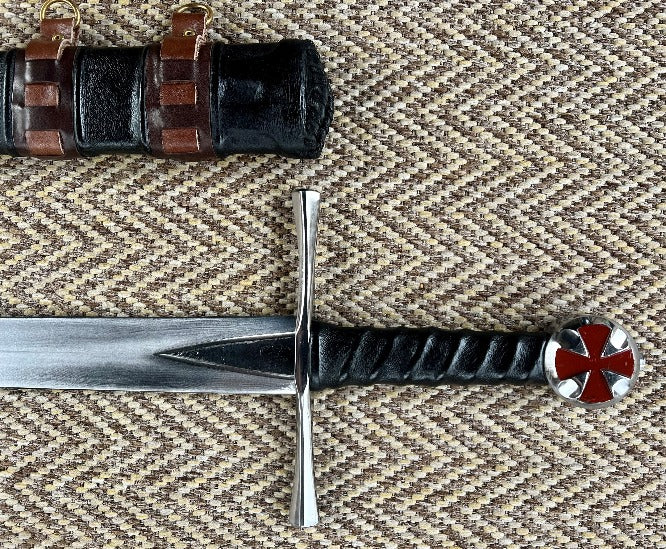 A close-up of a hand forged carbon steel blade with a leather wrapped handle, featuring a red cross inset in the pommel and a polished quillons. A wooden scabbard with leather accents is also visible in the background.