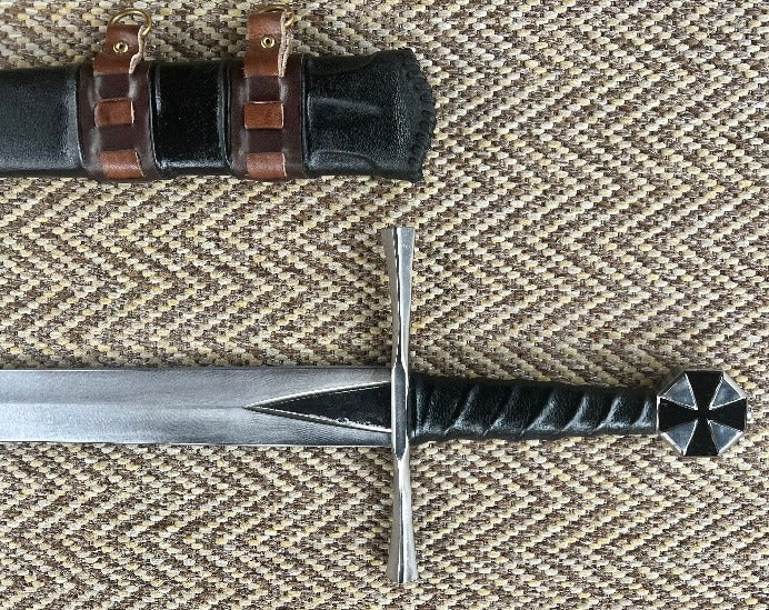 A close-up of a hand forged carbon steel blade with a black cross design, featuring a leather wrapped handle and a wooden scabbard with leather accents in the background.