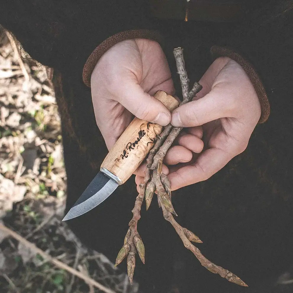 Roselli R130 The Grandmother Knife for craft work held in hands with branches outdoors.